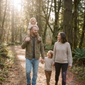 A father walking a sunlit woodland trail with his partner and two young children, one on his shoulders